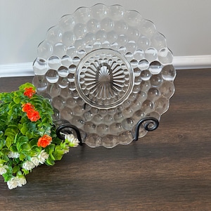 May include: A clear glass plate with a bubble design and a scalloped edge. The plate is displayed on a black metal stand. The plate has a central, raised, circular design. The plate is approximately 10 inches in diameter.