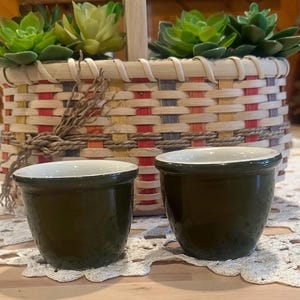 May include: Two small, dark green ceramic pots with white rims sit in front of a woven basket filled with faux succulents. The basket has a handle and is decorated with colorful woven patterns.