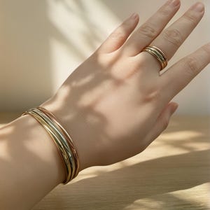 May include: A hand wearing a gold ring and a gold, rose gold, and yellow gold bangle bracelet set. The jewelry is displayed on a light wooden surface, with shadows cast across the hand and surface.