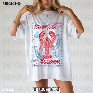 Op de afbeelding: Wit T-shirt met een afbeelding van een rode kreeft in een blauwe en roze schelp met de tekst "Crawfish Season" in een blauwe en roze rand.