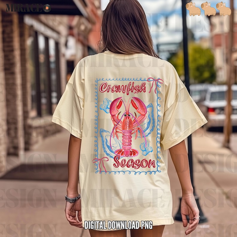 Op de afbeelding: Een lichtgeel T-shirt met een afbeelding van een roze kreeft met blauwe en witte accenten. De tekst "Crawfish Season" is in blauw en wit geschreven.