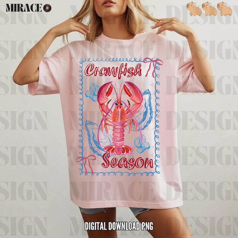 Op de afbeelding: Een roze T-shirt met een afbeelding van een kreeft in een blauwe en witte schelp met de tekst "Crawfish Season" in een blauwe en witte rand.