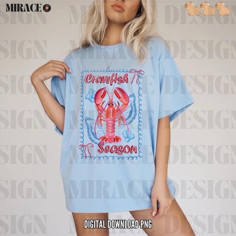 Op de afbeelding: Een lichtblauw T-shirt met een afbeelding van een rode kreeft in een blauw-witte schelp. De tekst "Crawfish Season" is onder de kreeft gedrukt.