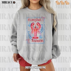 Op de afbeelding: Een grijs sweatshirt met een afbeelding van een rode kreeft met de tekst "Crawfish Season" in een blauwe en roze rand.