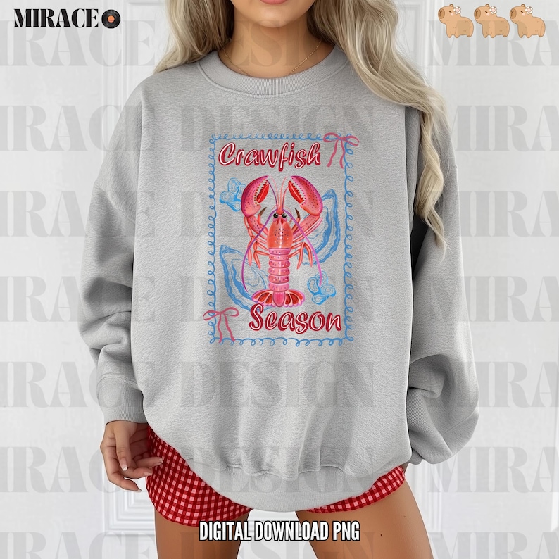 Op de afbeelding: Een grijs sweatshirt met een afbeelding van een rode kreeft met de tekst "Crawfish Season" in een blauwe en roze rand.