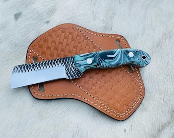 Puede incluir: Un cuchillo pequeño con mango negro y verde y hoja dentada. El cuchillo está apoyado en una funda de cuero marrón.