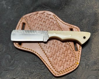 Puede incluir: Un pequeño cuchillo de acero inoxidable con mango de hueso blanco y una lima en la hoja. El cuchillo está en una funda de cuero marrón con un patrón tejido.