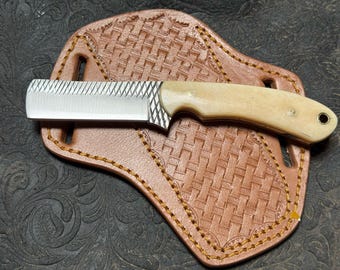 Puede incluir: Un pequeño cuchillo hecho a mano con mango de hueso y hoja dentada descansa sobre una funda de cuero marrón. La funda tiene un patrón tejido y está cosida con hilo amarillo. El cuchillo es una herramienta.