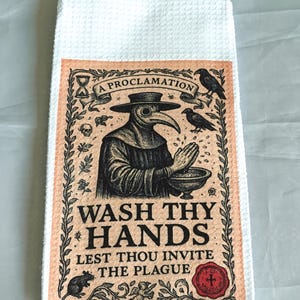 May include: White kitchen towel with a rectangular design featuring a vintage illustration of a plague doctor. The design includes the text "A Proclamation" and "Wash Thy Hands Lest Thou Invite The Plague."