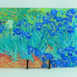 May include: Rectangular glass cutting board featuring a vibrant floral painting. The artwork depicts blue irises with green leaves and yellow flowers, reminiscent of a famous painting. The board is supported by a black metal stand.
