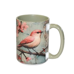 May include: A ceramic mug with a sage green interior and handle, featuring a printed illustration of a pink bird perched on a dark brown branch. The background displays a light blue textured surface with pink cherry blossom flowers and buds.