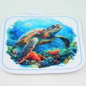 May include: Square white pot holder with a blue border and a loop for hanging. The center features a colorful illustration of a sea turtle swimming above coral reefs in the ocean. The turtle has orange, green, and brown markings.