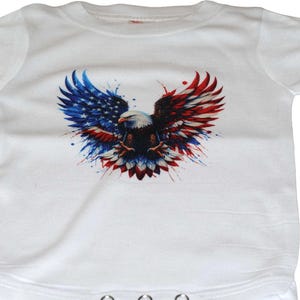 May include: White baby bodysuit featuring a patriotic design. The graphic depicts an eagle with wings spread, incorporating the colors of the American flag: red, white, and blue. The eagle's wings are stylized with a watercolor effect.