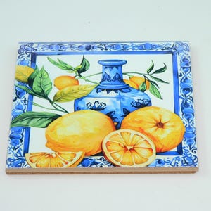 May include: Square ceramic tile with a painted design featuring a blue and white vase, lemons, and orange slices. The tile has a blue border with a decorative pattern and a cork backing.