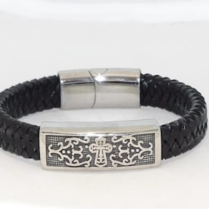 May include: Black braided leather bracelet with a stainless steel clasp. The bracelet features a decorative silver-tone rectangular clasp with an ornate cross design. A stylish accessory.