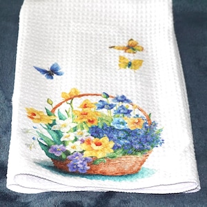May include: White waffle-weave kitchen towel featuring a watercolor design of a basket overflowing with yellow, blue, and purple flowers. Two yellow butterflies and one blue butterfly are also depicted on the towel.