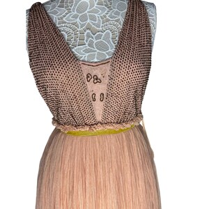 May include: A sleeveless, floor-length dress in shades of peach and tan. The bodice features a V-neck design with a beaded overlay. A thin, yellow-green belt cinches the waist, and the skirt is pleated. The dress is displayed on a mannequin.
