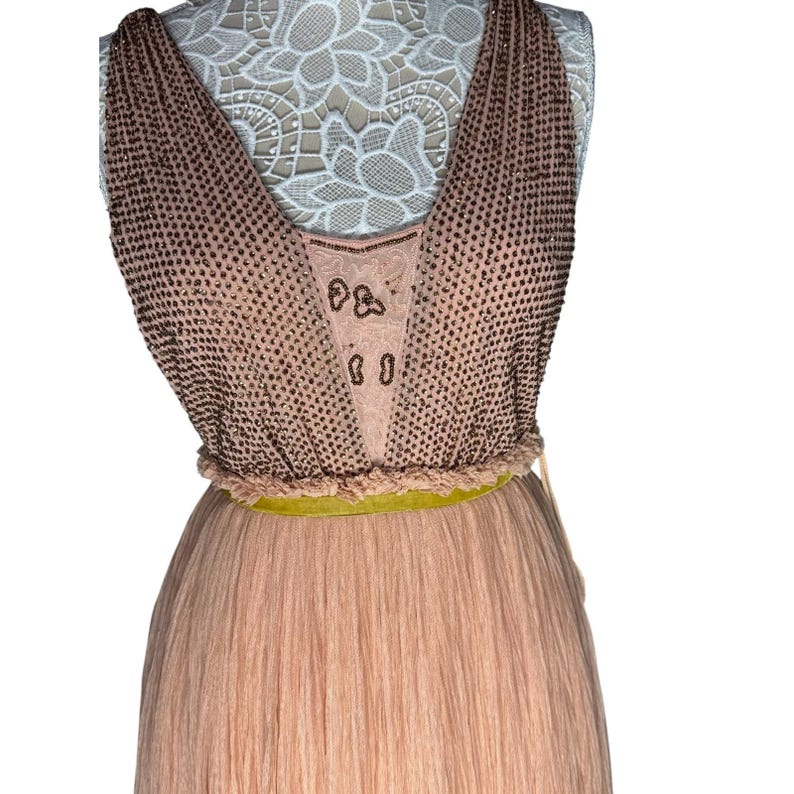 May include: A sleeveless, floor-length dress in shades of peach and tan. The bodice features a V-neck design with a beaded overlay. A thin, yellow-green belt cinches the waist, and the skirt is pleated. The dress is displayed on a mannequin.