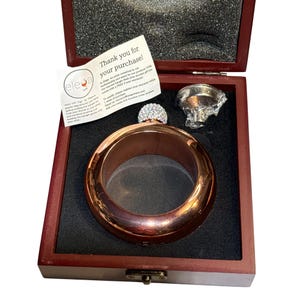 May include: A copper-coloured bangle flask with a crystal-studded cap and a silver-coloured funnel, presented in a dark wooden box. The box contains a thank you note. The flask is a stylish accessory.