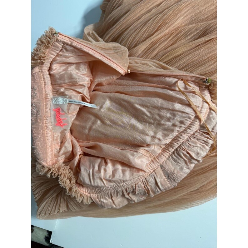 May include: A peach-colored dress with a layered, pleated skirt and a ruffled bodice. The interior lining is a lighter peach color, and the dress features a zipper closure. The label reads "Free People".