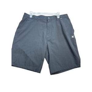 Puede incluir: Pantalones cortos de color gris oscuro con cierre de botón y logotipo blanco en la pierna. Los pantalones cortos cuelgan de una percha blanca. Los pantalones cortos tienen dos bolsillos delanteros y dos traseros.