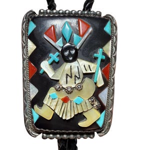 May include: A bolo tie with a rectangular, black centerpiece featuring a Native American figure inlaid with turquoise, red, and mother-of-pearl accents. The silver-toned frame has a beaded edge, and the tie has a black leather cord.