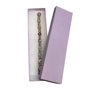 May include: A silver bracelet with colorful flower-shaped gemstones is displayed inside a light purple box. The bracelet features a series of flower-shaped gems in various colors, including yellow, red, and clear. The box is open, revealing the bracelet against a white interior.