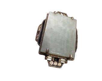 Pode incluir: Anel retangular de jade com moldura prateada. O jade é de cor verde pálido. O anel tem um design decorativo com pequenas pedras transparentes e detalhes pretos.