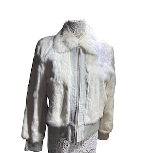 Könnte beinhalten: Eine weiße Kunstfell-Bomberjacke mit silbernem Reißverschluss. Die Jacke hat einen gerippten Stricksaum und Bündchen sowie eine weiche, flauschige Textur. Der Kragen ist ebenfalls mit Kunstfell verziert.