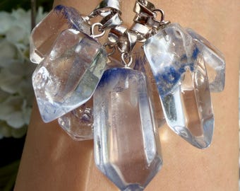 Op de afbeelding: Een cluster van heldere, puntige kristalachtige hangers die aan een zilverkleurige metalen ketting hangen. Elke hanger heeft een opvallende blauwgetinte insluiting aan de basis, met een glad, gefacetteerd oppervlak dat het licht opvangt.