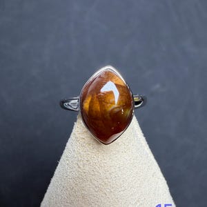 Op de afbeelding: Een zilveren ring met een marquise-vormige, gepolijste bruine edelsteen met amberkleurige wervelingen en donkere insluitingen. De steen is gezet in een gladde metalen zetting en het getal 15 staat in blauw rechtsonder in de afbeelding.