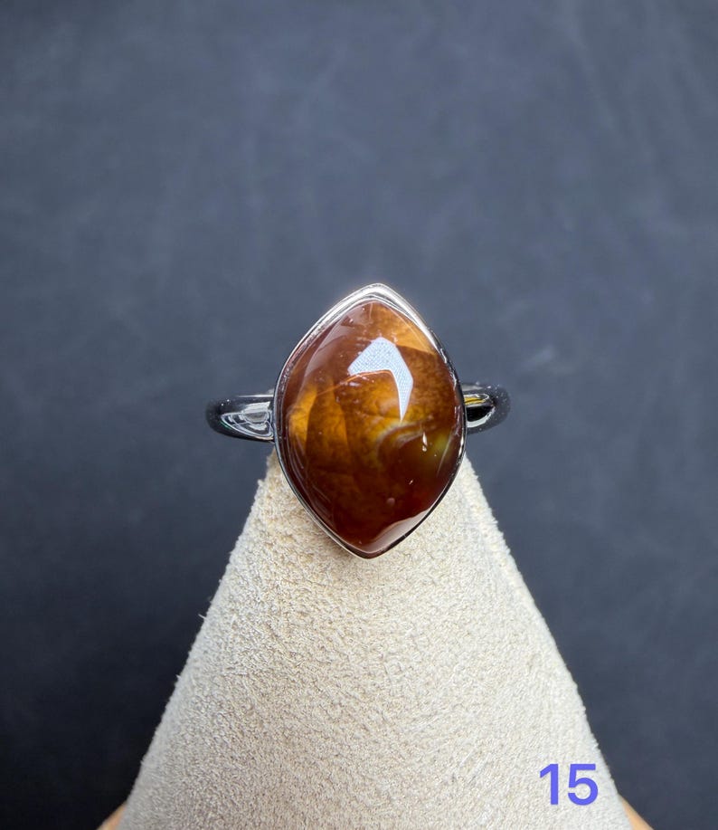 Op de afbeelding: Een zilveren ring met een marquise-vormige, gepolijste bruine edelsteen met amberkleurige wervelingen en donkere insluitingen. De steen is gezet in een gladde metalen zetting en het getal 15 staat in blauw rechtsonder in de afbeelding.