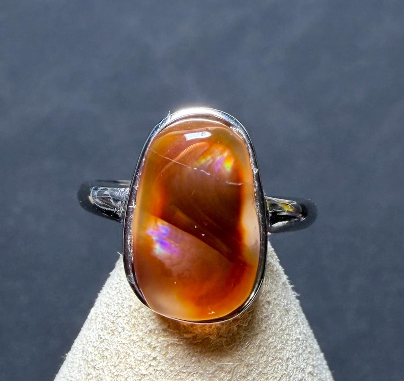 Op de afbeelding: Een zilverkleurige ring met een grote, gladde, ovaalvormige cabochonsteen. De steen vertoont een diepe, doorschijnende oranje en roodbruine kleur met interne iriserende lichtflitsen in paars, blauw en geel door de steen heen.