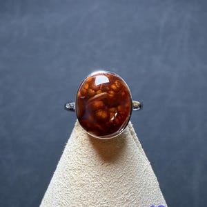Op de afbeelding: Een zilveren ring met een grote, ovaalvormige cabochonsteen in een diepe roodbruine kleur met een opvallend, bubbelachtig of orbiculair intern patroon. De steen is gezet in een gladde, gepolijste zilveren zetting met een dunne ringband.