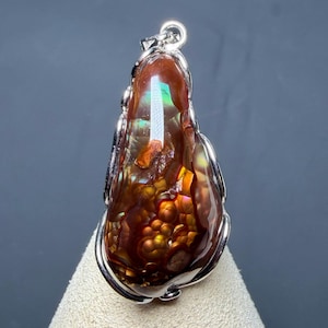 May include: A fire agate pendant set in a polished silver frame. The gemstone features a teardrop shape with deep reddish-brown tones, displaying iridescent flashes of green, gold, and orange light across its textured, botryoidal surface.