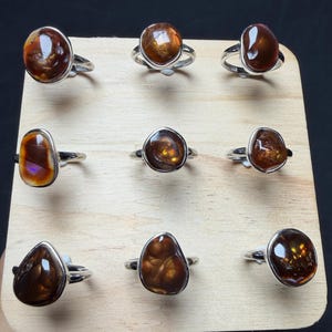 Op de afbeelding: Een collectie van negen zilveren ringen, elk voorzien van een unieke, gepolijste, onregelmatig gevormde bruine vuuragaat edelsteen. De ringen zijn gerangschikt in een drie-bij-drie raster op een licht houten displaybord, met diverse tinten amber en bruin.