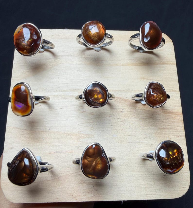 Op de afbeelding: Een collectie van negen zilveren ringen, elk voorzien van een unieke, gepolijste, onregelmatig gevormde bruine vuuragaat edelsteen. De ringen zijn gerangschikt in een drie-bij-drie raster op een licht houten displaybord, met diverse tinten amber en bruin.