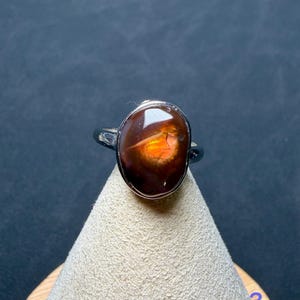 Op de afbeelding: Een zilveren ring met een gepolijste, ovaalvormige vuuragaat edelsteen. De steen heeft een diepbruine, doorschijnende basis met een levendig, gloeiend oranje en geel iriserend centrum. De metalen ringband is glad en reflecterend.
