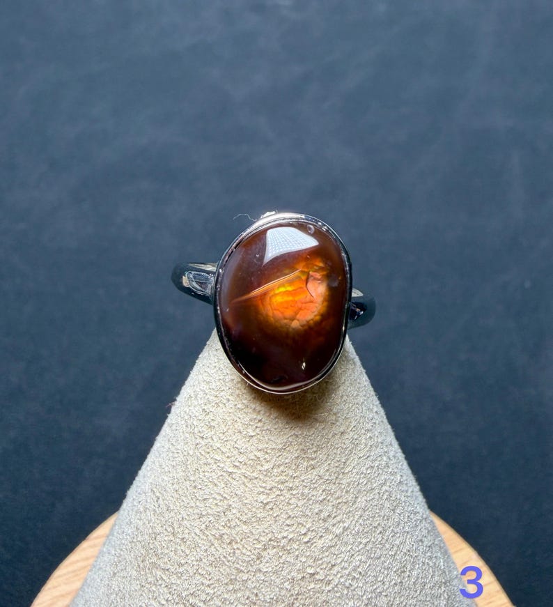 Op de afbeelding: Een zilveren ring met een gepolijste, ovaalvormige vuuragaat edelsteen. De steen heeft een diepbruine, doorschijnende basis met een levendig, gloeiend oranje en geel iriserend centrum. De metalen ringband is glad en reflecterend.