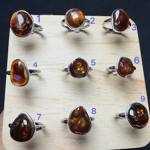 Op de afbeelding: Negen zilveren ringen met gepolijste, onregelmatige, bruine vuuragaat edelstenen, elk gemarkeerd met een blauw cijfer van 1 tot 9. De ringen zijn in een raster op een lichtgekleurde houten plank gerangschikt en tonen diverse vormen.