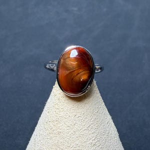 Op de afbeelding: Een zilverkleurige ring met een grote, ovaalvormige cabochonsteen met een wervelend patroon in dieproodbruin en metaalachtig koper. De steen is gezet in een gladde, gepolijste metalen zetting en de ringband is van eenvoudig, effen zilverkleurig metaal.