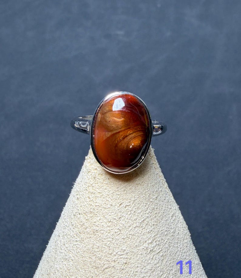 Op de afbeelding: Een zilverkleurige ring met een grote, ovaalvormige cabochonsteen met een wervelend patroon in dieproodbruin en metaalachtig koper. De steen is gezet in een gladde, gepolijste metalen zetting en de ringband is van eenvoudig, effen zilverkleurig metaal.