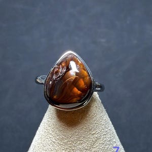 Op de afbeelding: Een zilveren ring met een druppelvormige vuuragaat edelsteen met diepbruine en oranje wervelende patronen. De gepolijste metalen band houdt de steen vast in een gladde zetting, wat de natuurlijke glans en kleurvariaties benadrukt.
