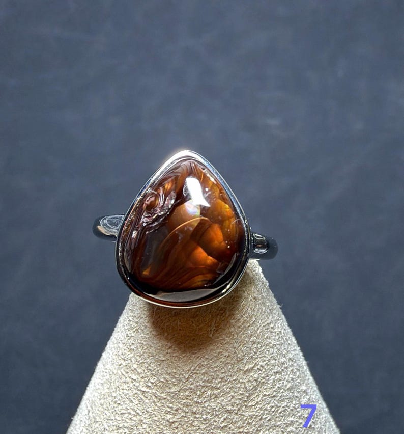 Op de afbeelding: Een zilveren ring met een druppelvormige vuuragaat edelsteen met diepbruine en oranje wervelende patronen. De gepolijste metalen band houdt de steen vast in een gladde zetting, wat de natuurlijke glans en kleurvariaties benadrukt.
