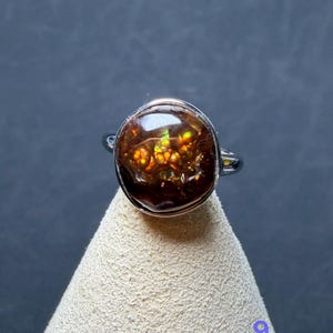 Op de afbeelding: Een zilverkleurige ring met een ronde, gepolijste vuuragaat edelsteen. De steen is donkerbruin met interne flitsen van levendig oranje, geel en groen licht. De metalen band is glad en gepolijst en omsluit de cabochon geslepen steen.