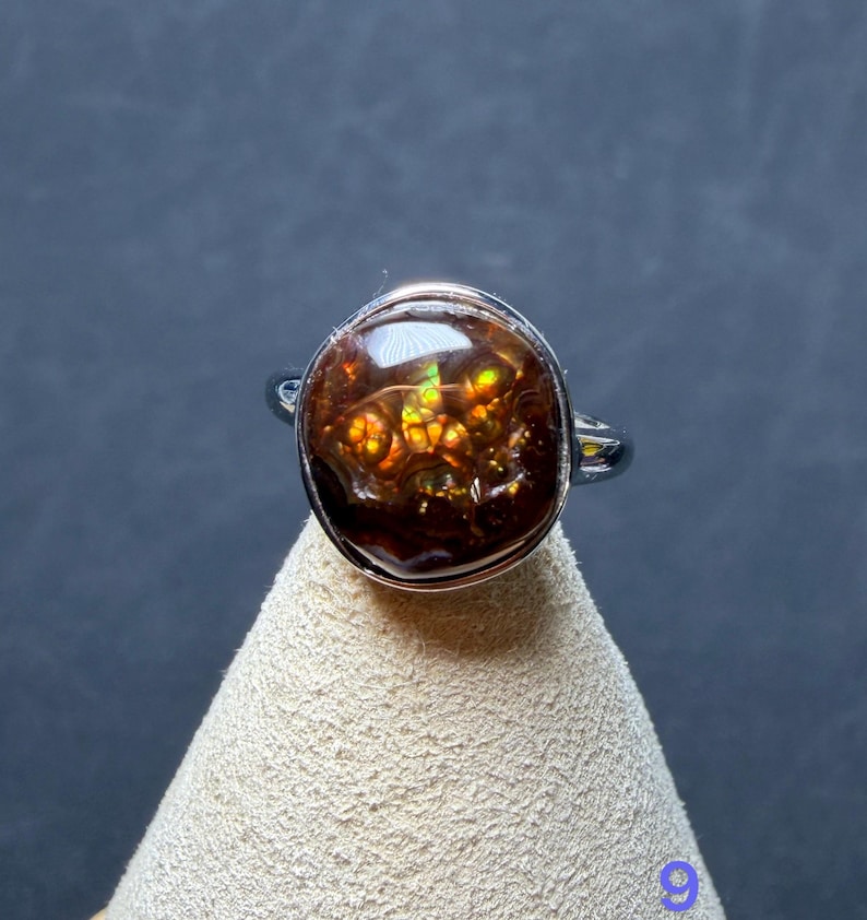 Op de afbeelding: Een zilverkleurige ring met een ronde, gepolijste vuuragaat edelsteen. De steen is donkerbruin met interne flitsen van levendig oranje, geel en groen licht. De metalen band is glad en gepolijst en omsluit de cabochon geslepen steen.