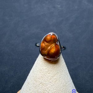 Op de afbeelding: Een zilveren ring met een gepolijste, onregelmatige, druppelvormige vuuragaat edelsteen met donkeroranje, bruine en roodbruine wervelende patronen. De steen is gezet in een gladde metalen zetting, het cijfer 8 is zichtbaar in de hoek.