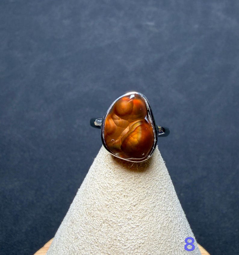Op de afbeelding: Een zilveren ring met een gepolijste, onregelmatige, druppelvormige vuuragaat edelsteen met donkeroranje, bruine en roodbruine wervelende patronen. De steen is gezet in een gladde metalen zetting, het cijfer 8 is zichtbaar in de hoek.