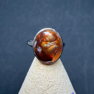 Op de afbeelding: Een zilveren ring met een grote, ovaalvormige vuuragaat edelsteen. De steen heeft een diepe roodbruine basiskleur met iriserende flitsen van oranje, goud en groen licht die reflecteren vanuit het gepolijste, afgeronde oppervlak.