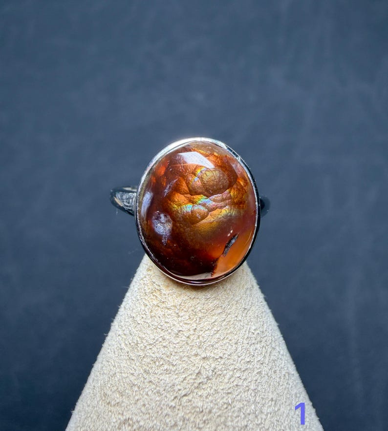 Op de afbeelding: Een zilveren ring met een grote, ovaalvormige vuuragaat edelsteen. De steen heeft een diepe roodbruine basiskleur met iriserende flitsen van oranje, goud en groen licht die reflecteren vanuit het gepolijste, afgeronde oppervlak.